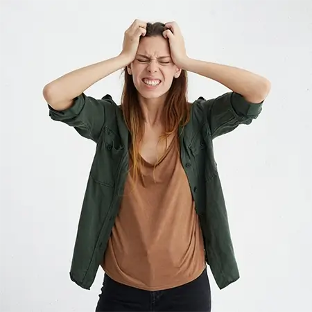 imagem de uma mulher, de camisa marrom com outra de botão, verde, por cima, com as mãos na cabeça, em sinal de irritação