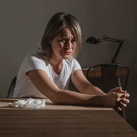 mulher sentada em uma mesa com um expressão de mal estar no rosto, de camisa branca, com as mãos sobre a mesa
