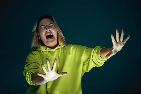 mulher de casaco verde florescente com expressão de pânico no rosto, fazendo sinal com as mãos para afastar algo