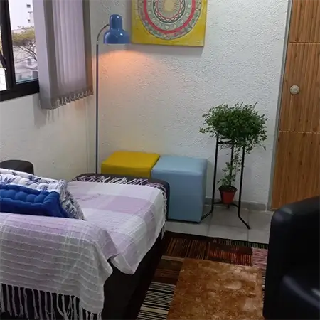 imagem da sala de atendimento com um sofá preto com uma colcha listrada, lilás e branco, com 2 almofadas, com 2 puffs no canto, um amarelo e outro azul, um vaso de plantas e um tapete quadriculado ao centro