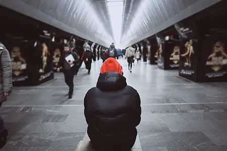 pessoa de casaco preto e gorro laranja sentada no chão da estação de trem, com outras pessoas passando ao redor