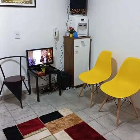 imagem da recepção, com duas cadeiras amarelas lado a lado, perto de um bebedouro e de uma mesa com uma tv, com um tapete quadriculado no centro