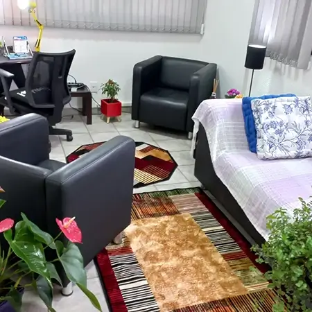 imagem do interior da sala de atendimento, com um tapete no centro, sofás individuais pretos, um sofá maior preto coberto com uma colcha listrada, lilás e branco, 2 almofadas e uma mesa de computador ao fundo, preta com uma cadeira preta