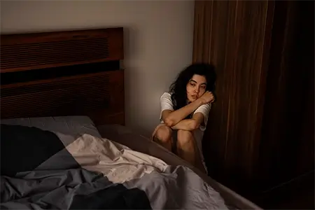 imagem com uma mulher encolhida no canto do quarto, de camisa branca, agarrada nas pernas, entre a cama e o armário