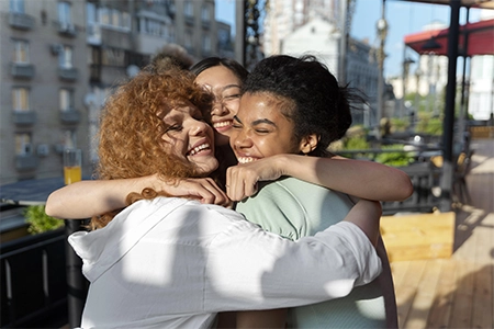 três amigas se abraçando e sorrindo, com a cidade, ao fundo